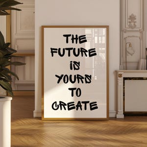 Könnte beinhalten: Ein gerahmter Druck mit dem Text "THE FUTURE IS YOURS TO CREATE" in schwarzer Pinselbeschriftung. Der Rahmen ist goldfarben, und der Druck wird vor einem weißen Hintergrund angezeigt. Das Kunstwerk lehnt an einer weißen Wand in einem Raum mit Parkettboden.