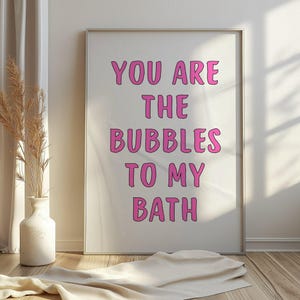 Puede incluir: Una impresión enmarcada con el texto "YOU ARE THE BUBBLES TO MY BATH" en letras rosas. La impresión se muestra en una pared blanca, con un jarrón de plantas secas y un paño doblado en primer plano.