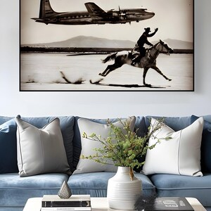 May include: A rectangular wall art print in a black frame featuring a sepia-toned photograph of a person riding a horse across a flat landscape while a propeller airplane flies overhead. The scene includes distant mountains under a clear sky.
