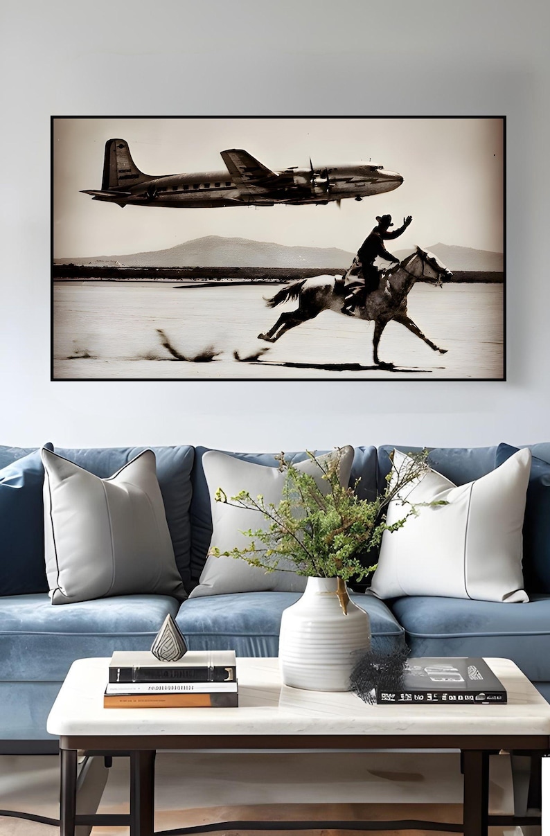 May include: A rectangular wall art print in a black frame featuring a sepia-toned photograph of a person riding a horse across a flat landscape while a propeller airplane flies overhead. The scene includes distant mountains under a clear sky.