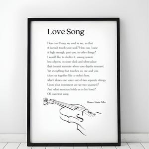 May include: Black framed print of the poem "Love Song" by Rainer Maria Rilke. The text is in black on a white background, with a violin illustration. The print is displayed on a white wooden surface.