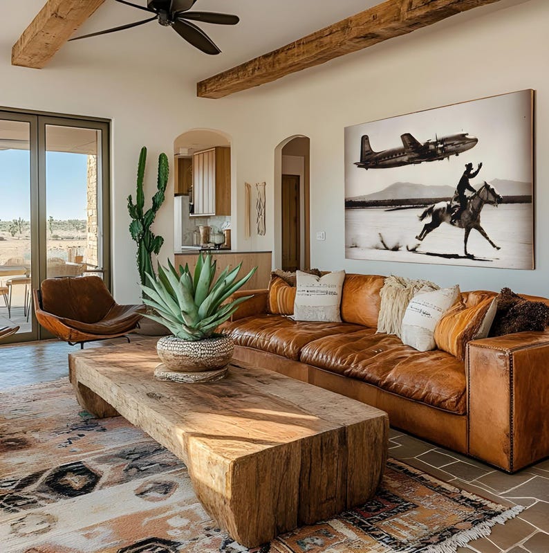 May include: A large living room featuring a brown leather sofa with decorative pillows, a low rectangular coffee table made from a solid piece of natural wood, and a large black and white wall art print depicting a horse rider and an airplane.