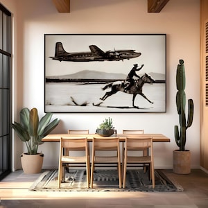 May include: A large rectangular black and white wall art print featuring a vintage airplane flying over a desert landscape where a person is riding a horse at a gallop. The artwork is framed in a thin black border and displayed on a white wall.