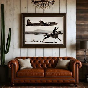 May include: A framed black and white wall art print depicting a vintage airplane flying above a person riding a horse across a flat landscape. The artwork is displayed above a brown leather tufted sofa with two light-colored throw pillows.