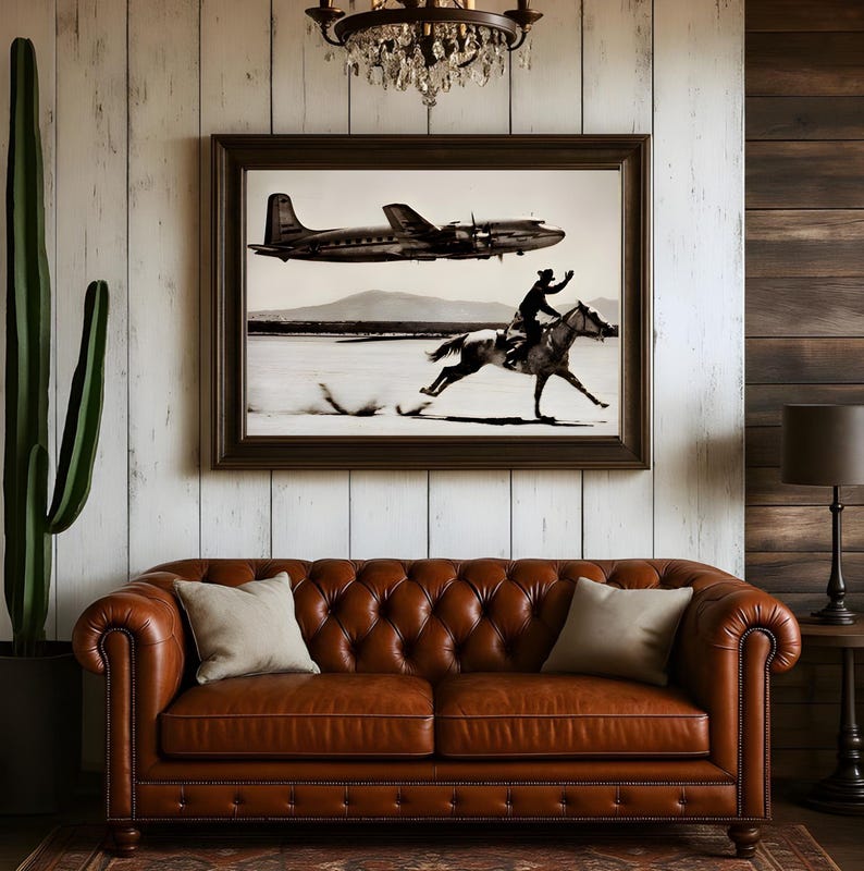 May include: A framed black and white wall art print depicting a vintage airplane flying above a person riding a horse across a flat landscape. The artwork is displayed above a brown leather tufted sofa with two light-colored throw pillows.