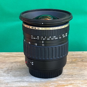 May include: A black and gold camera lens with the text "Di II (IF) 11-18mm 1:4.5-5.6" and measurement markings. The lens is cylindrical with a black lens hood and sits on a wooden surface against a green backdrop.