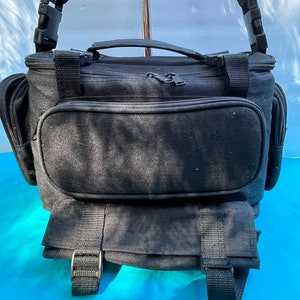 May include: Black camera bag with multiple compartments and adjustable shoulder strap. Features a top handle, front pocket, and side pockets. The bag has a buckle closure and is made of a durable, textured material. Ideal for photographers.