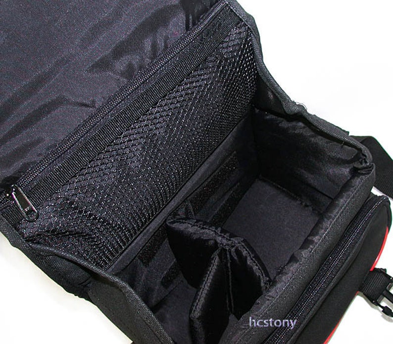 May include: A black camera bag with an open top, featuring a zippered mesh pocket on the underside of the lid, a main compartment with adjustable padded dividers, and the text hcstony printed in small white letters on the lower right side.