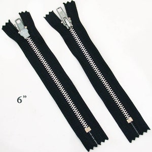 May include: Two black 6-inch zippers with silver teeth and pulls. The zippers are laid out side by side against a white background. The measurement "6" is printed in black on the left side.