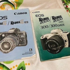 May include: Two instruction manuals for Canon EOS Rebel cameras. The left manual is for the Rebel G and Rebel G QD cameras. The right manual is for the Rebel 2000 and Rebel 2000 300/300 DATE cameras. Both manuals feature a photo of the camera on the cover.