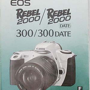 May include: A white Canon EOS Rebel 2000/2000 Date 300/300 Date camera with a Canon Zoom Lens EF 28-80mm 1:4-5.6 attached. The camera has a black lens cap and a black strap.