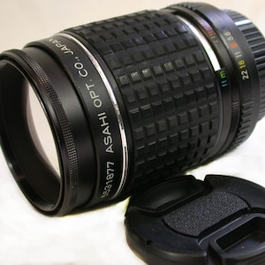 May include: A black camera lens with a silver band that reads "5531877 ASAHI OPT. CO., JAPAN". The lens has a depth of field scale marked in feet and meters. The lens is accompanied by a black lens cap.