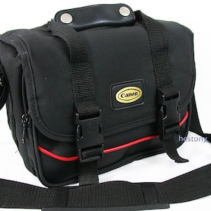 May include: A black camera bag featuring a top handle with silver rivets, two adjustable black nylon straps with plastic side-release buckles, and a red piping accent. A gold and black Canon logo patch is centred on the front flap.