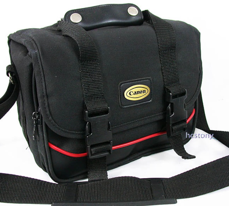 May include: A black camera bag featuring a top handle with silver rivets, two adjustable black nylon straps with plastic side-release buckles, and a red piping accent. A gold and black Canon logo patch is centred on the front flap.