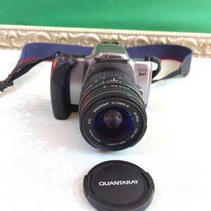 May include: A Canon EOS Rebel XS camera with a Quantaray lens and lens cap. The camera is silver and black with a black lens. The lens cap is black with white text. The camera strap is blue with red accents.