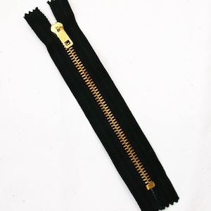 May include: A black zipper with a gold-colored metal zipper pull and teeth. The zipper is open and angled across a white background. The zipper teeth are a contrasting gold color against the black fabric.
