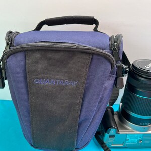 May include: A navy blue Quantaray camera bag with a black front panel and a black handle. A Canon camera with a lens is next to the bag. The lens has a black cap.