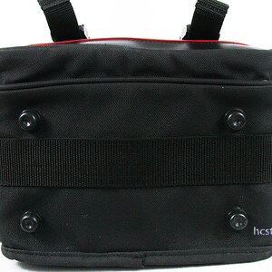 May include: A black rectangular bag featuring a textured fabric exterior, a wide horizontal strap across the centre, and four circular black feet on the bottom panel. A thin red accent line is visible along the top edge, and the text hcstony is printed in the bottom right corner.