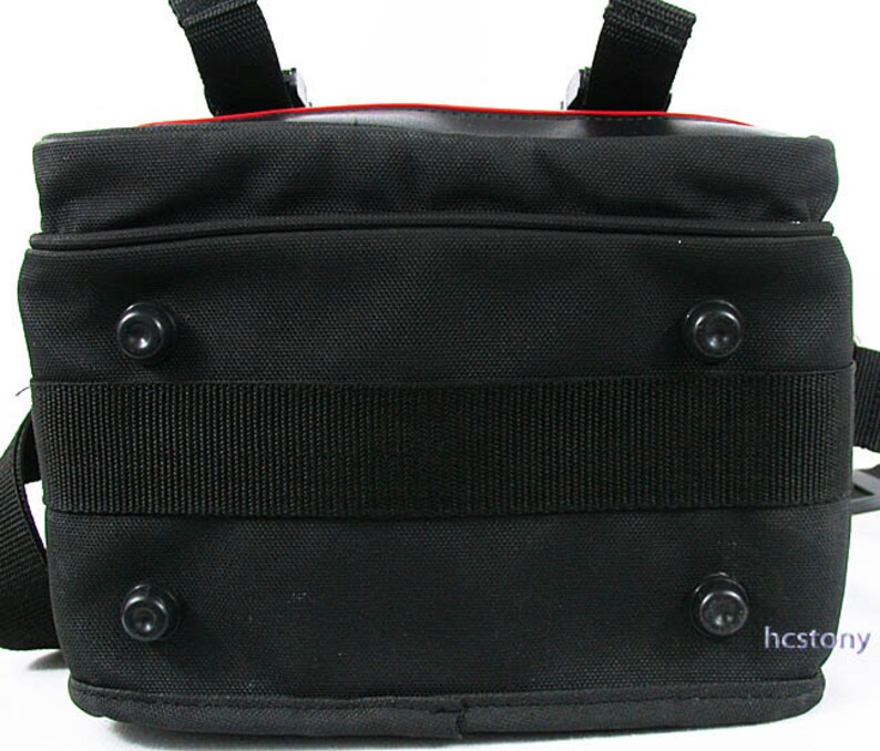 May include: A black rectangular bag featuring a textured fabric exterior, a wide horizontal strap across the centre, and four circular black feet on the bottom panel. A thin red accent line is visible along the top edge, and the text hcstony is printed in the bottom right corner.