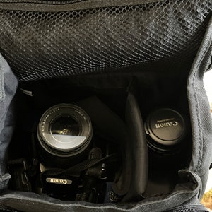 May include: A black camera bag containing a camera with an EF-S 18-55mm 1:3.5-5.6 lens attached and a separate lens with a Canon Ultrasonic cap. The bag features a zipped top compartment with a black mesh pocket and internal padded dividers.