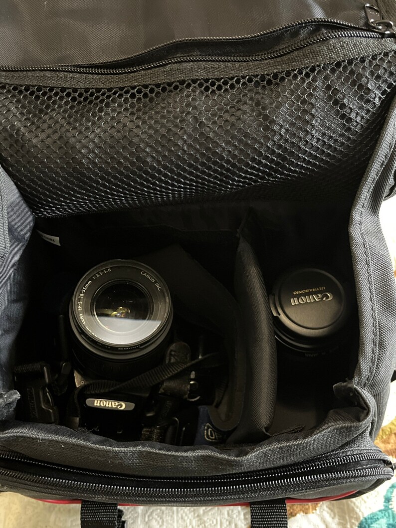 May include: A black camera bag containing a camera with an EF-S 18-55mm 1:3.5-5.6 lens attached and a separate lens with a Canon Ultrasonic cap. The bag features a zipped top compartment with a black mesh pocket and internal padded dividers.