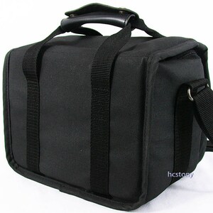 May include: A black rectangular camera bag made of durable fabric with a top handle featuring a padded grip. The bag includes two vertical black webbing straps on the front, a side attachment point for a shoulder strap, and the text hcstony in blue.