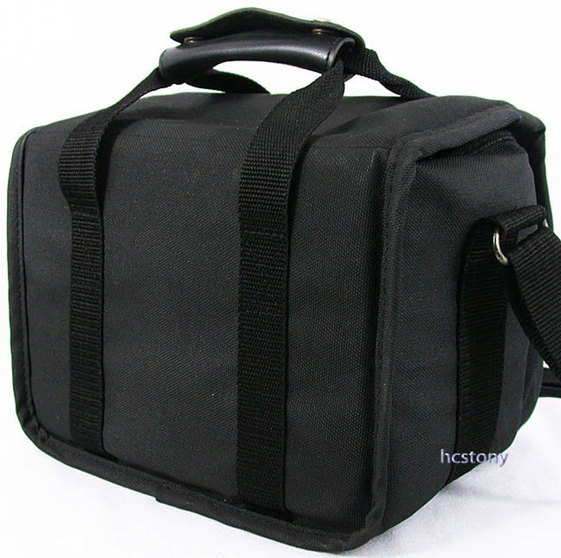 May include: A black rectangular camera bag made of durable fabric with a top handle featuring a padded grip. The bag includes two vertical black webbing straps on the front, a side attachment point for a shoulder strap, and the text hcstony in blue.