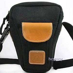 May include: Black camera bag with a tan leather flap and a smaller leather patch. The bag has a shoulder strap and a buckle closure. The text "hcstony" is visible on the lower right side of the bag.