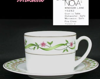 May include: A white Mikasa Studio Nova Windsor Lane Y0282 teacup and saucer set. The teacup has a gold rim and a floral design with pink flowers and green leaves. The saucer has a matching floral pattern.