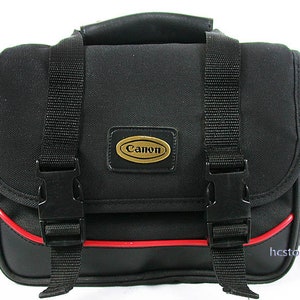 May include: A black camera bag featuring a top handle, two black plastic side-release buckles on the front flap, and a red horizontal trim accent. An oval-shaped gold and black badge with the text Canon is centred on the front of the bag.