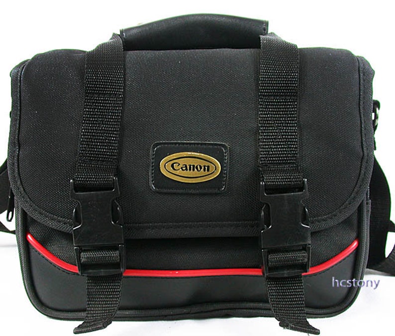 May include: A black camera bag featuring a top handle, two black plastic side-release buckles on the front flap, and a red horizontal trim accent. An oval-shaped gold and black badge with the text Canon is centred on the front of the bag.