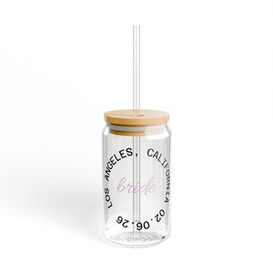 Puede incluir: Un vaso de vidrio transparente con tapa y pajita de bambú. El vaso tiene las palabras "LOS ANGELES, CALIFORNIA" en un círculo alrededor de la palabra "bride" en escritura rosa claro. La fecha "02.06.90" está impresa debajo.