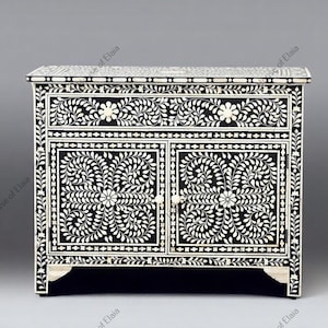 May include: Black and white floral inlaid cabinet with two doors and a drawer. The cabinet features intricate leaf and flower patterns. The cabinet has a rectangular shape and is made of wood. The cabinet has a decorative design and is a piece of furniture.