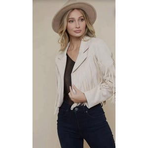 Può includere: Giacca in camoscio beige con frange sulle maniche e sul davanti. La giacca ha un taglio blazer classico ed è abbinata a un cappello coordinato. L'outfit è completato da jeans blu scuro.