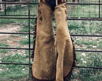 May include: Brown leather chaps with decorative diamond shapes and fringe along the sides. The chaps are worn over blue jeans and have a brown leather belt with a silver buckle. The chaps are designed for horseback riding.