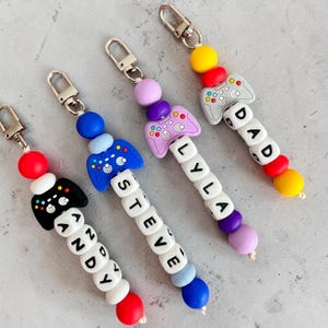 May include: Four colorful keychains with game controller charms. Each keychain features a different color scheme, with names spelled out in white letter beads. The names are Andy, Steve, Lyla, and Dad.