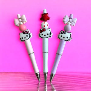 May include: Three Hello Kitty pens with different colored tops. The pen on the left has a pink top with a bow and beads. The middle pen has a white top with a red star and red and white polka dot beads. The pen on the right has a purple top with a bow and beads.