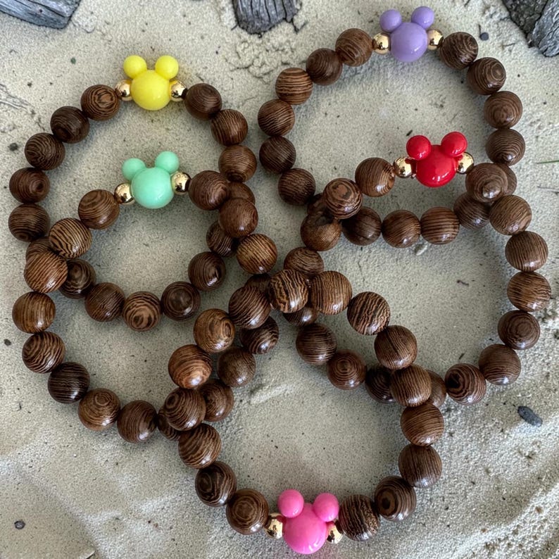 May include: Four wooden bead bracelets with colorful Mickey Mouse-shaped accents. The bracelets feature brown wooden beads and Mickey Mouse heads in yellow, mint green, purple, and pink. The bracelets are on a sandy surface.