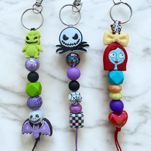 May include: Three Nightmare Before Christmas-themed keychains. Each keychain features a different character: Oogie Boogie, Jack Skellington, and Sally. The keychains are made of colorful beads and charms, with silver-tone metal clasps.