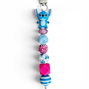 May include: A beaded keychain featuring a blue character charm at the top, followed by a sequence of beads including a translucent pink bead, a light blue faceted bead, a bright pink rhinestone bead, a white bead with blue floral patterns, a hot pink cube, and a blue and white striped sphere.