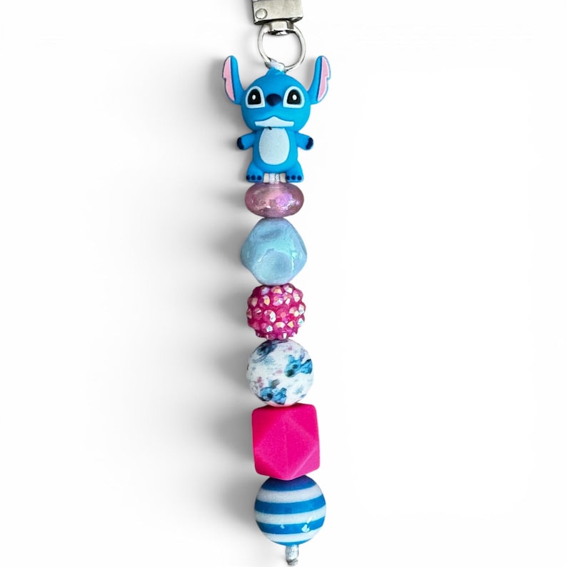 May include: A beaded keychain featuring a blue character charm at the top, followed by a sequence of beads including a translucent pink bead, a light blue faceted bead, a bright pink rhinestone bead, a white bead with blue floral patterns, a hot pink cube, and a blue and white striped sphere.