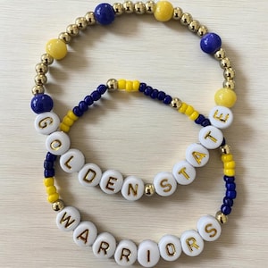 May include: Two beaded bracelets. One bracelet spells out "GOLDEN STATE" in white letter beads with gold lettering, and the other spells out "WARRIORS". The bracelets feature blue, yellow, and gold beads.