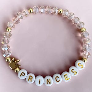 May include: A beaded bracelet with clear, faceted beads and gold-colored accents. The bracelet features white letter beads spelling "PRINCESS" and a gold-colored crown charm. The background is a solid pink color.