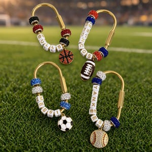 May include: Four gold-toned metal carabiner keychains featuring white letter beads spelling BULLS, BRONCOS, MESSI, and YANKEES. Each keychain includes colorful crystal-encrusted beads and a sports-themed charm: a basketball, football, soccer ball, or baseball.