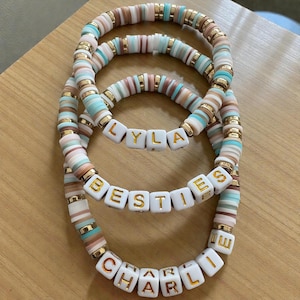 May include: Three stacked bracelets with colorful, flat beads in shades of pink, blue, brown, and gold. The bracelets spell out "LYLA", "BESTIES", and "CHARLIE" with white lettered beads.