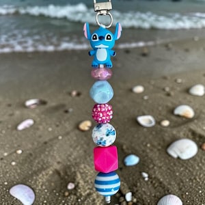 May include: A beaded keychain featuring a blue cartoon character charm at the top, followed by a sequence of beads including a translucent pink bead, a light blue faceted bead, a bright pink rhinestone bead, a floral patterned bead, a hot pink cube, and a blue and white striped bead.