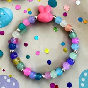 May include: A colorful beaded bracelet featuring a pink bunny charm. The bracelet is made of various colored beads, including pink, blue, green, and purple, separated by silver-colored beads. A fun and playful accessory.