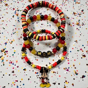 Puede incluir: Tres pulseras de cuentas de colores y un collar. Las pulseras presentan cuentas rojas, negras y amarillas. El collar tiene un dije de Mickey Mouse y la palabra "MOM" escrita con cuentas negras.