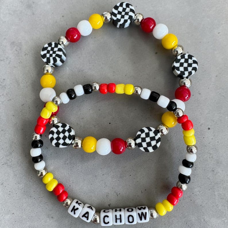 May include: Two beaded bracelets with a racing theme. The bracelets feature red, yellow, white, black, and silver beads. Some beads have a checkered pattern. One bracelet spells out "KACHOW" with white letter beads.
