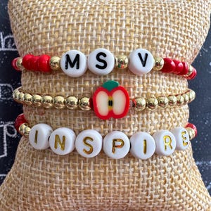 May include: Three beaded bracelets. The top bracelet has white letter beads spelling "MSV" between red beads. The middle bracelet has gold beads and a red apple charm. The bottom bracelet has white letter beads spelling "INSPIRE".